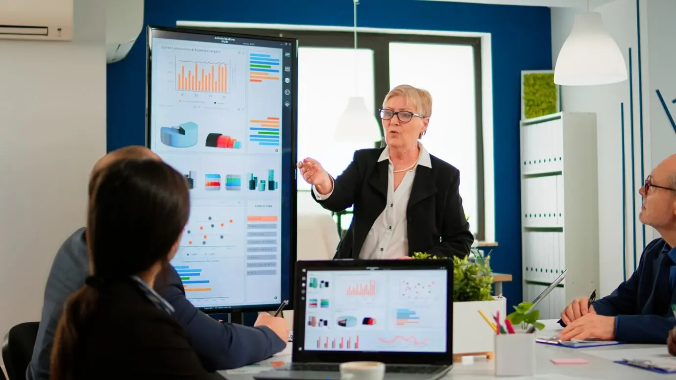 Пожилой менеджер проекта, указывая на настольный компьютер, представляет статистические данные и проводит брифинг для разнообразной группы сотрудников. Многоэтническая команда работает в профессиональной финансовой компании-стартапе во время конференции.